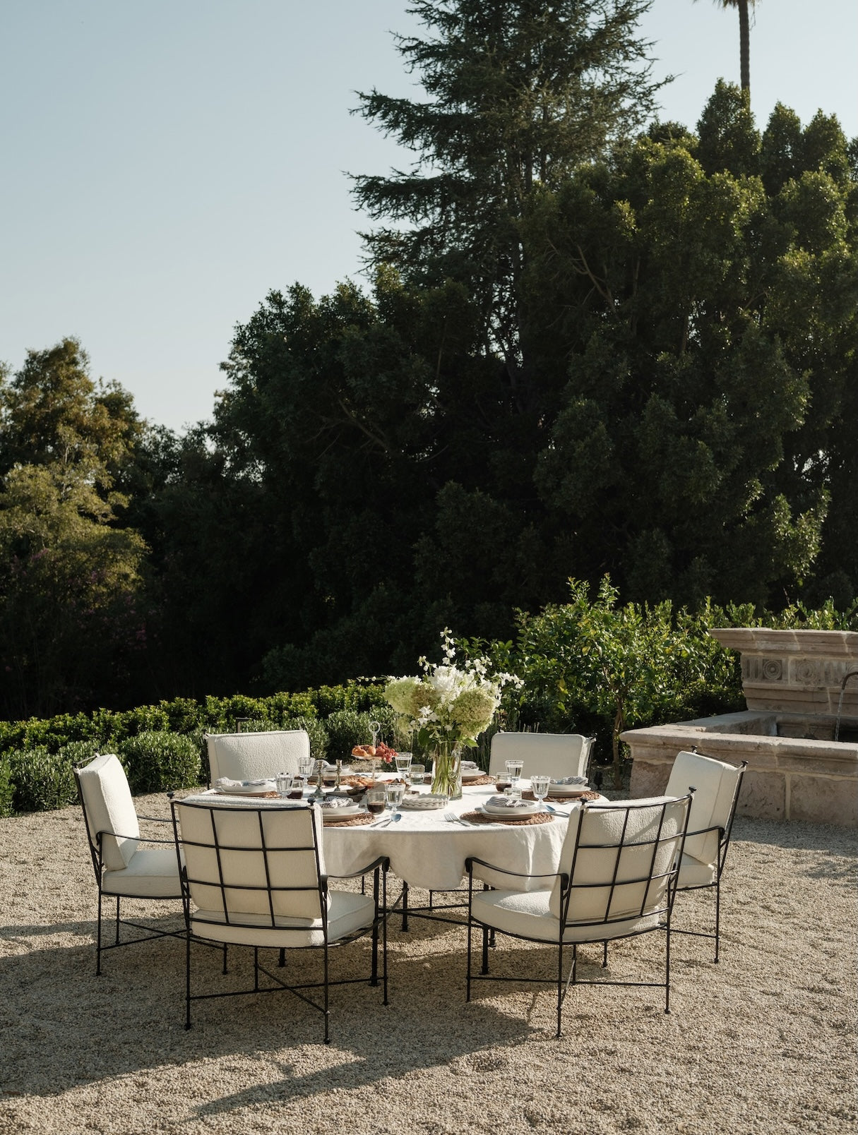 Main Image A stylish outdoor dining setting with a round table, white chairs, surrounded by lush greenery under a clear sky.
