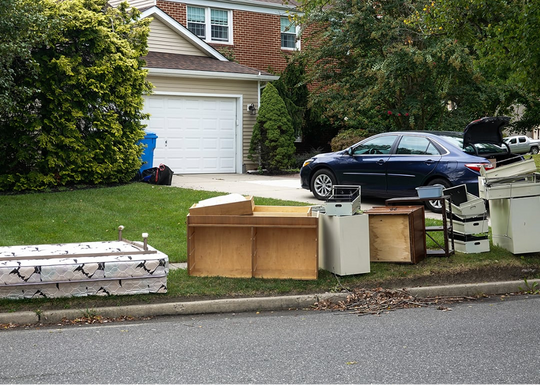 Old furniture and items placed on the curb in front of a suburban home, with a car in the driveway.