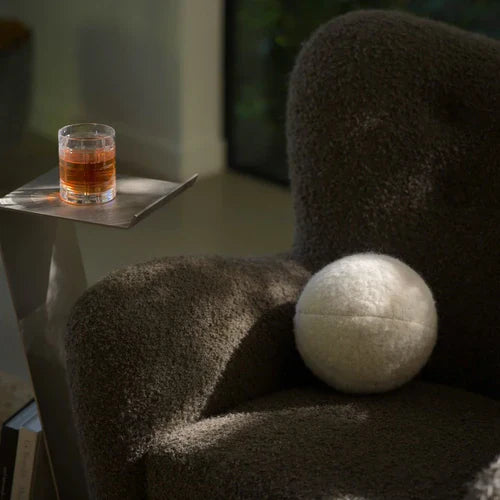 Plush gray armchair with a white round pillow, next to a sleek metal side table holding a glass of drink.
