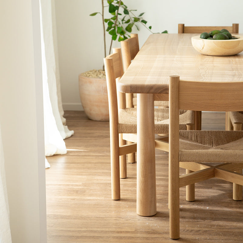 Minimalist wooden dining table with matching chairs, a bowl of fruit, and a potted plant in a bright.
