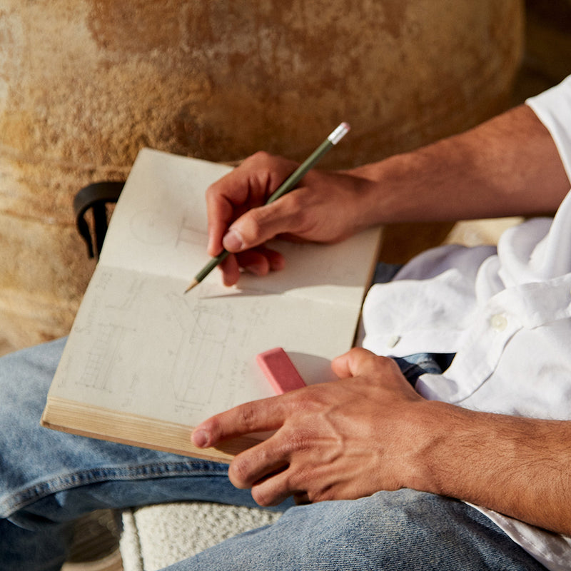 Person sketching in a notebook with a pencil, holding an eraser, wearing a white shirt and jeans, seated outdoors.
