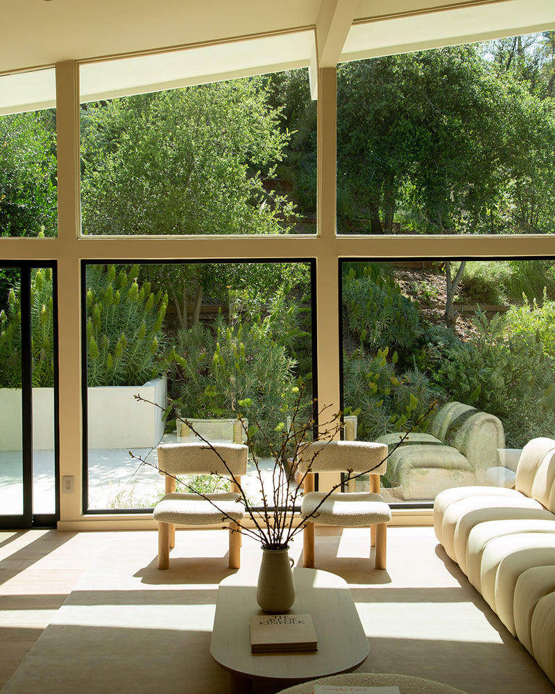 Bright living room with floor-to-ceiling windows, modern furniture, and a view of lush greenery in the background.
