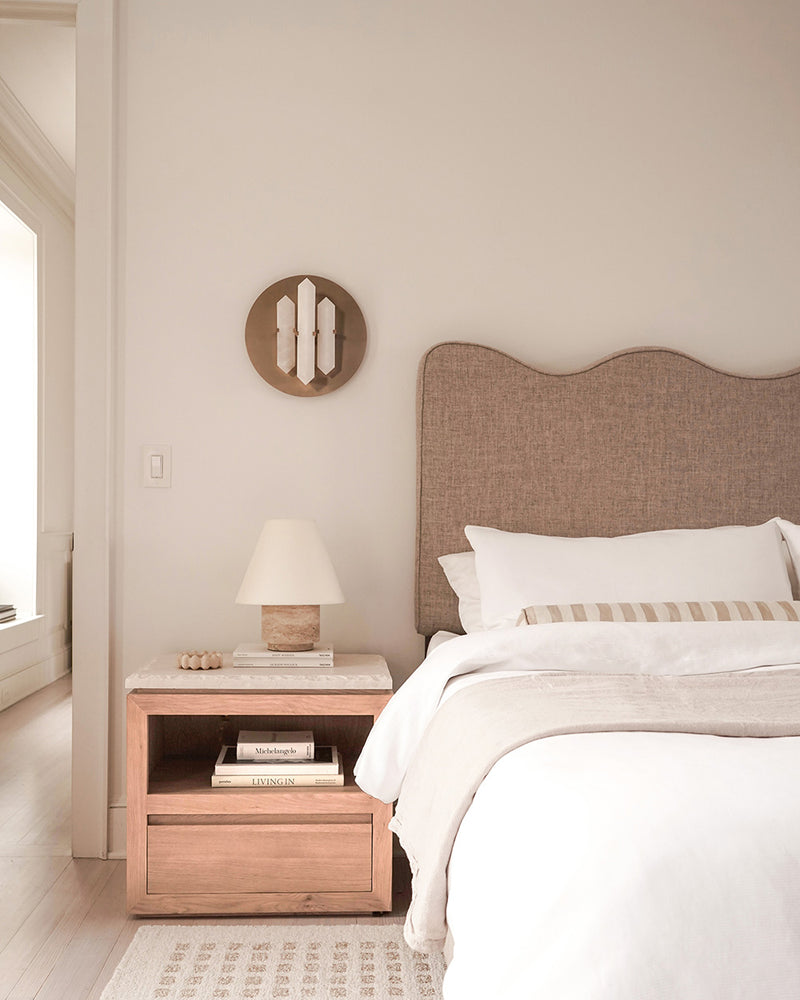 Minimalist bedroom with a beige upholstered headboard, wooden nightstand, table lamp, and neutral bedding in soft lighting.